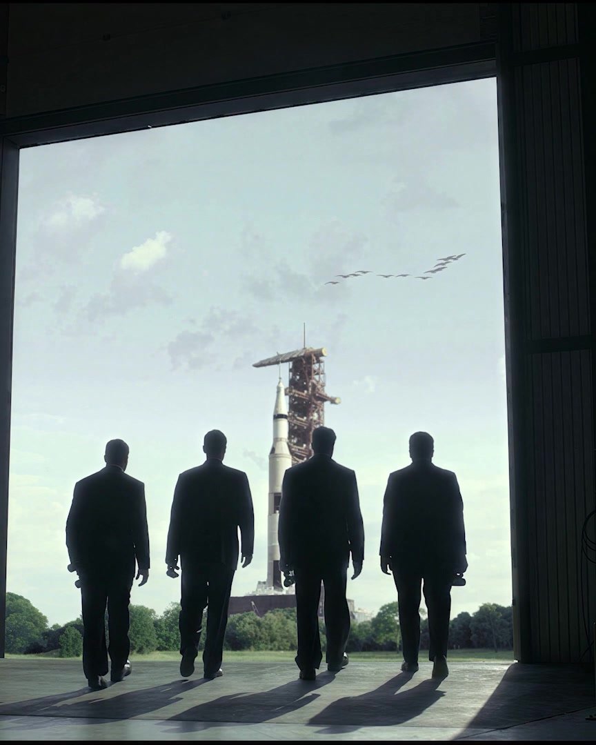 Four silhouetted figures walking out of a dark hangar toward a rocket on the pad